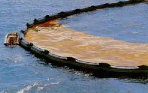 A photo showing part of a large, black rubber barrier which contains a brown oil spill floating on blue sea water. There is a small boat pushing the black rubber barrier inwards to concentrate the oil spill.