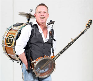 A man carrying a drum and cymbals on his back, a banjo in front and a holder which holds a harmonica and microphone to his mouth.
