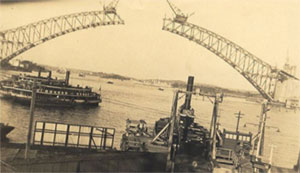 Photo showing a half built Sydney Harbour Bridge shown across Sydney Harbour. The upper arch is in 2 parts and has not yet met in the middle. The roadway has not yet been added. The triangles that make up the arch are clearly visible. 