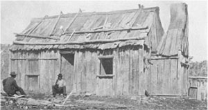 A photo showing a rough hut made of planks of wood. The hut is rectangular in shape with a sloping roof. There are 2 windows, one of which is open, the other is shut. The door is open and a man is sitting on the small step leading to the front door. Another man is sitting on some logs, facing the man in the doorway.  Both men are wearing hats. There is a large box-like structure on the side of the house. It has a chimney, made out of the same type of wood attached to the top of the structure. This structure probably contains the kitchen/cooking area of the house.