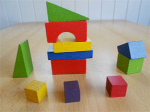 11 different coloured, different shaped, wooden building blocks. 5 are stacked forming a small tower and others are scattered around its base.