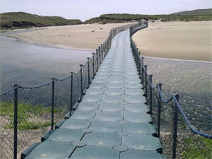 A bridge which floats on the water, crossing from one bank of a river to another. The bridge consists of a path that people can walk on, a hand rail and safety mesh on both sides of the pathway running along the length of the pathway.