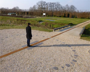A person is standing in the centre of a large circle, flat on the ground,  in a park in daylight. The edge of the circle has flat stones placed at equal distances around it. There is a straight line going through the centre of the circle, extended through the top of the circle and the bottom of the circle. The person is standing up straight on the line going through the circle. The person’s shadow extends from the centre of the circle to the edge of the circle and beyond.